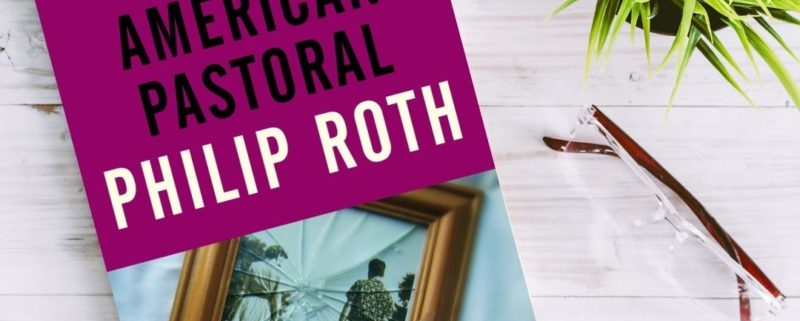 A copy of American Pastoral by Philip Roth on a white wooden table next to a plant and a pair of reading glasses.