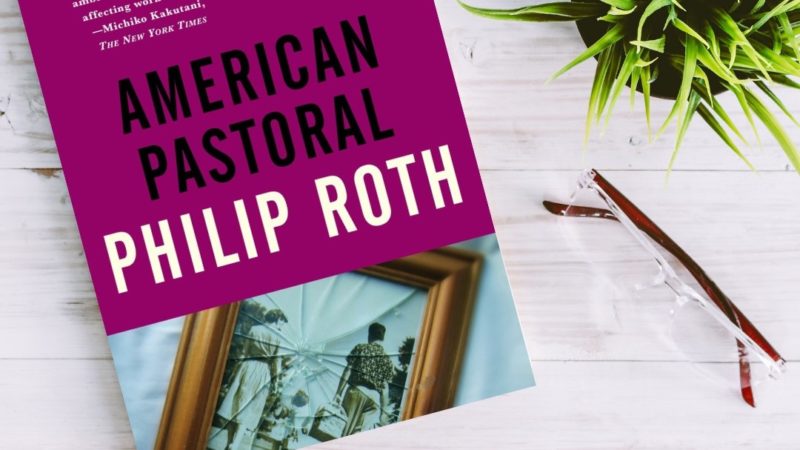 A copy of American Pastoral by Philip Roth on a white wooden table next to a plant and a pair of reading glasses.